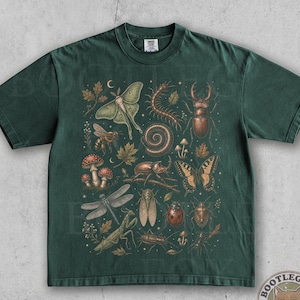 May include: A dark green short-sleeve t-shirt featuring a detailed graphic print of various insects, including a moth, beetle, dragonfly, ladybug, and centipede, interspersed with small mushrooms, leaves, and stars across the center chest area.