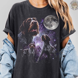 Peut inclure: Un t-shirt gris anthracite avec une impression graphique représentant trois chiens d'arrêt allemands à poil dur sur un fond de nébuleuse violette avec une pleine lune. Le motif inclut une grande tête de chien, un chien debout et un plus petit.