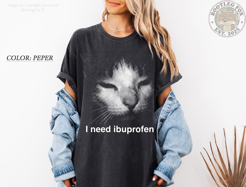 May include: A dark gray t-shirt with a distressed cat face graphic. The text "I need ibuprofen" is printed below the cat's face. The shirt is a pepper color.