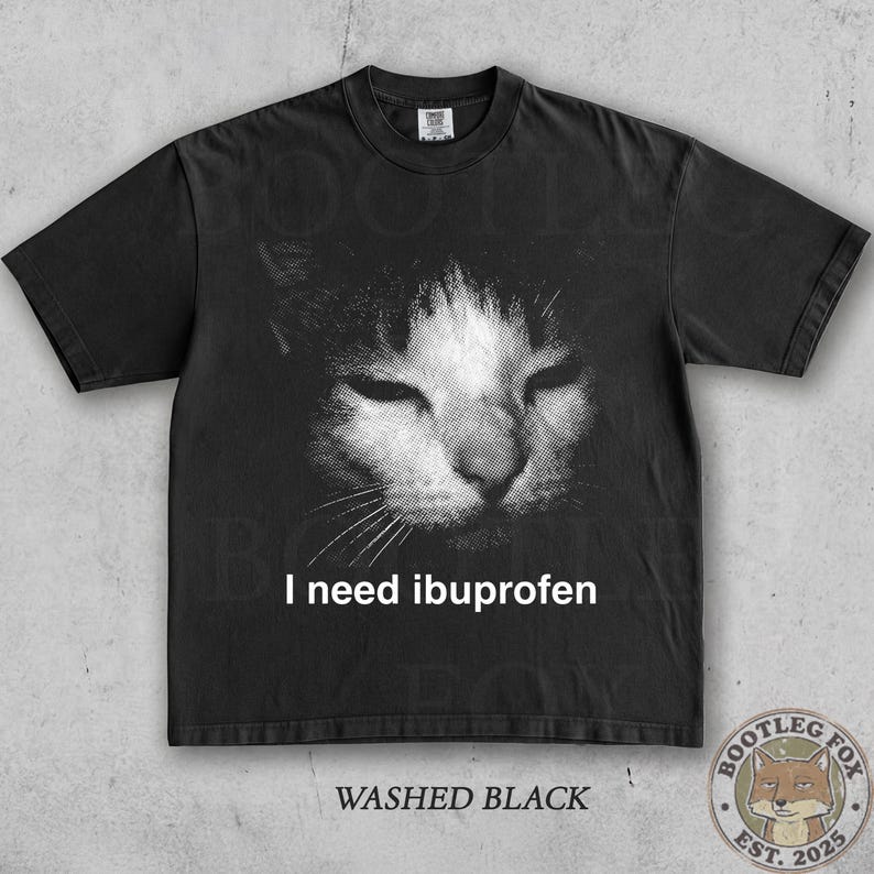 May include: Black t-shirt with a distressed graphic of a cat's face in black and white. The text "I need ibuprofen" is printed below the cat image. The shirt is labeled "Washed Black".