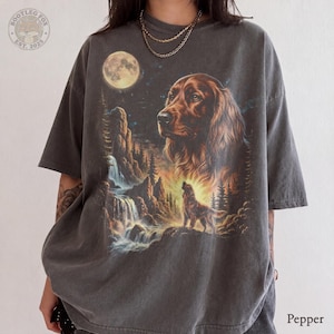 May include: A charcoal gray t-shirt featuring a graphic print of a large brown dog head above a smaller dog standing near a waterfall and a full moon. The shirt includes a circular logo in the top left corner with the text Bootleg Fox Est. 2025.
