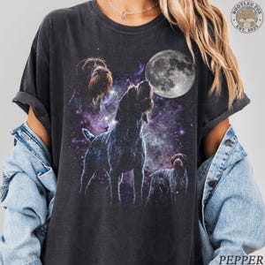 May include: Dark grey t-shirt with a graphic of three dogs set against a cosmic backdrop and a full moon. The dogs are in brown and black, the moon in grayscale. The shirt has the text "BOOTLEG FOX EST. 2025" and "PEPPER".
