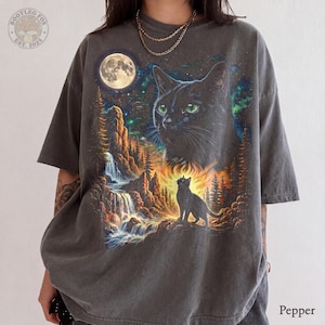 May include: A charcoal grey short-sleeve t-shirt featuring a graphic print of a large black cat head with green eyes, a smaller black cat silhouette, a waterfall, rocky cliffs, pine trees, a full moon, and a starry night sky.