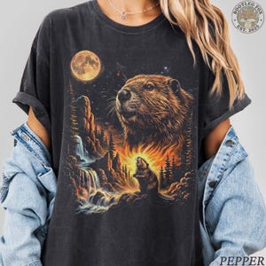 May include: Dark gray t-shirt featuring a detailed illustration of a beaver with a full moon and waterfall scene. The design includes a starry night sky and a smaller beaver silhouette. The shirt has a vintage, worn look.
