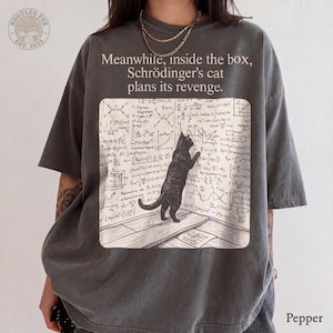 以下が含まれることがあります： チャコールグレーの半袖Tシャツ。木製の床に立ち、複雑な物理学の方程式で埋め尽くされた壁を見つめる黒猫のグラフィックが描かれています。テキストは「Meanwhile, inside the box, Schrödinger's cat plans its revenge.」と記されています。