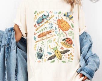 Puede incluir: Una camiseta de color marfil con un estampado gráfico colorido de varios insectos, incluyendo un escarabajo azul, un escarabajo amarillo anaranjado, una abeja, una mariquita y un ciervo volante, entre pequeñas hojas y ramas verdes.