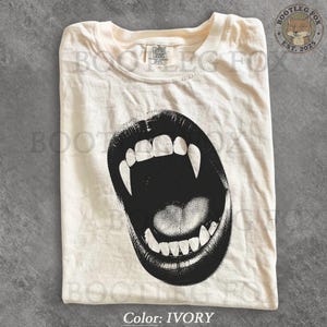 Könnte beinhalten: Ein elfenbeinfarbenes T-Shirt mit einem großen, schwarzen Grafikdruck eines offenen Mundes mit markanten Fangzähnen. Das Shirt hat einen Rundhalsausschnitt und ein Comfort Colors Markenetikett am inneren Halsausschnitt.