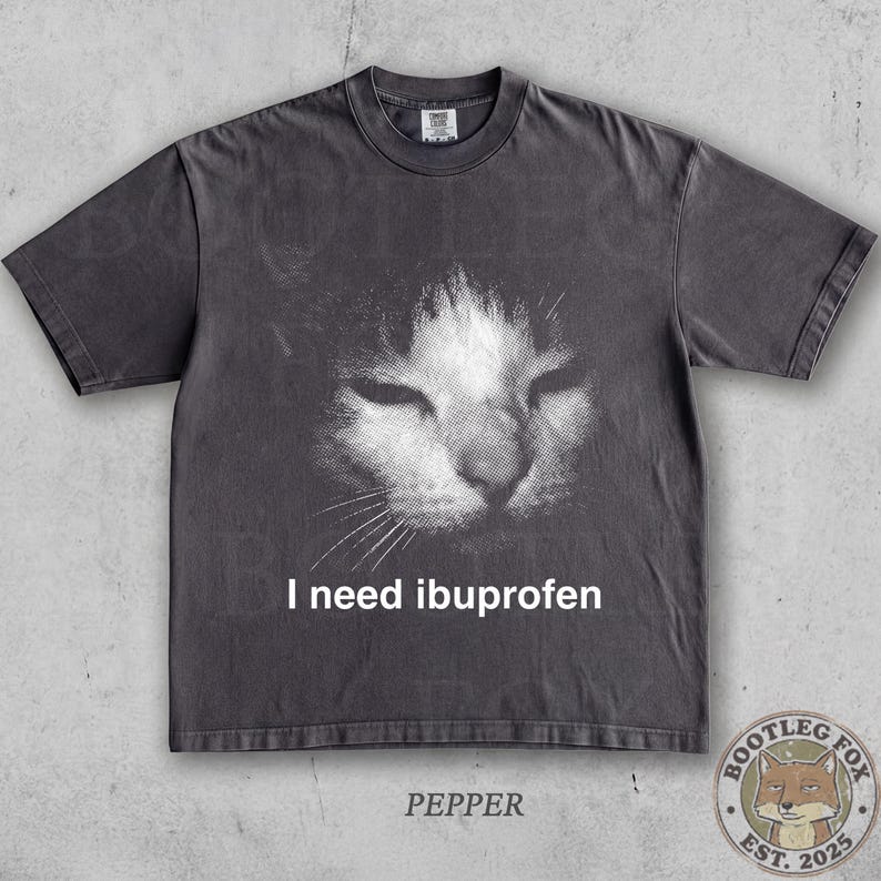 May include: Dark gray t-shirt featuring a grayscale image of a cat's face with closed eyes. The text "I need ibuprofen" is printed below the cat image. The word "PEPPER" is printed at the bottom.
