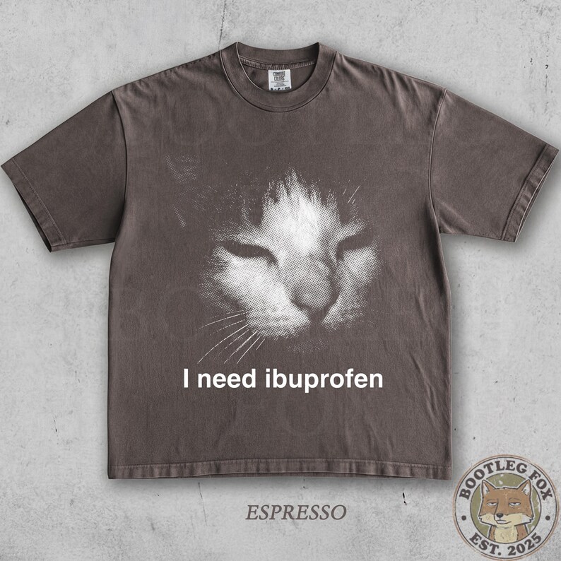 May include: Espresso-colored t-shirt featuring a distressed graphic of a cat's face with closed eyes. The text "I need ibuprofen" is printed below the image. A small logo with a fox is at the bottom.