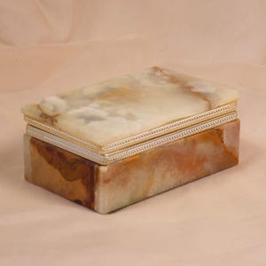 Peut inclure: Une boîte à bijoux rectangulaire avec un couvercle et une base en onyx marbré. Le couvercle est un mélange de crème, de beige et de gris, avec une bordure argentée. La base est un mélange de couleurs marron, beige et crème.