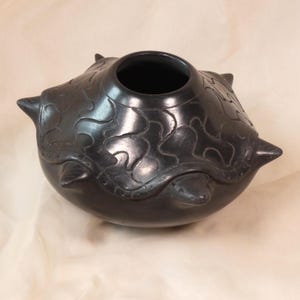 Può includere: Un vaso in ceramica grigio scuro con corpo arrotondato e ampia apertura. Il vaso presenta disegni stilizzati in rilievo e quattro elementi appuntiti sporgenti. La superficie ha una finitura opaca e la forma generale ricorda una tartaruga.