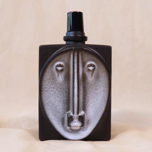 Può includere: Una bottiglia in ceramica marrone scuro con un design a volto stilizzato in bianco e grigio. La bottiglia ha una forma rettangolare con un tappo nero. Il volto presenta dettagli di occhi e naso.