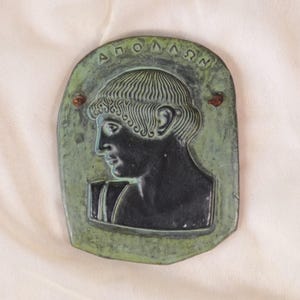 Op de afbeelding: Een bronzen plaquette met een groene patina, met een profiel van een man met krullend haar en de Griekse inscriptie "ΑΠΟΛΛΩΝ" bovenaan. Twee kleine, roodbruine accenten zijn zichtbaar. Rechthoekige vorm met afgeronde randen.