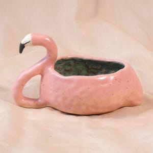 Peut inclure: Un pot de fleurs en céramique rose en forme de flamant rose avec un intérieur vert. Le flamant rose a un cou courbé, un bec noir et une pointe blanche. Un objet décoratif unique.