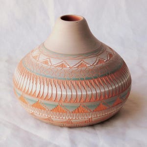 May include: A handmade pottery vase with a rounded body and a narrow neck. The vase displays intricate geometric patterns in orange, white, and teal against a beige background. The design includes triangles, lines, and textured areas.