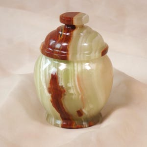 Op de afbeelding: Een decoratieve pot met deksel, gemaakt van onyx. De pot heeft een afgeronde body en een deksel met een kleine knop. De onyx toont een mix van groene, bruine en crèmekleurige kleuren, waardoor een natuurlijk, wervelend patroon ontstaat.