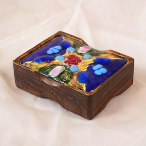 Puede incluir: Una caja de madera con una tapa colorida tipo mosaico. La tapa presenta un fondo azul oscuro con detalles azules, rosas y rojos dispersos. La caja tiene un acabado marrón oscuro.