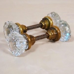 Peut inclure: Deux poignées de porte anciennes en verre avec des ferrures en laiton. Les boutons en verre transparent présentent un design à facettes, et les composants en laiton montrent des signes d'âge. Une tige métallique rouillée les relie.