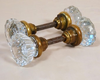 Op de afbeelding: Twee antieke glazen deurknoppen met messing hardware. De heldere glazen knoppen hebben een facetontwerp en de messing componenten vertonen tekenen van ouderdom. Ze zijn verbonden door een roestige metalen staaf.