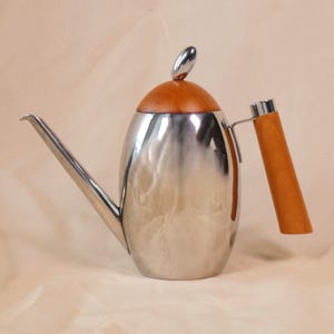 May include: A stainless steel gooseneck kettle with a wooden handle and lid. The kettle has a long, slender spout and a polished, reflective surface. The lid is made of wood and has a small, silver handle. The handle is made of wood and is attached to the kettle with silver hardware.