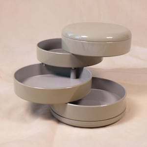 May include: A three-tiered, rotating storage container in a muted green-grey colour. The top tier is a lid, and the other two tiers are open trays. The container is designed to hold small items and is made of a smooth, glossy material.