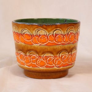 May include: A ceramic plant pot with a brown and orange pattern. The pot has a green interior and a pedestal base. The design features repeating orange circular shapes on a brown background. Suitable for indoor or outdoor use.