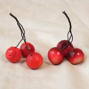 May include: Six red, round decorative cherries with dark brown twisted wire stems. The cherries have a glossy finish and varying shades of red. The cherries are grouped in pairs and singles.