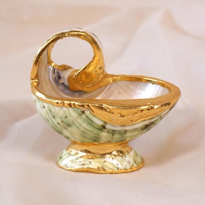Op de afbeelding: Een decoratieve schelpvormige kom met een handvat, met een groen en wit exterieur met gouden randen. De kom heeft een parelmoer interieur en staat op een kleine voet, eveneens afgewerkt met goud.