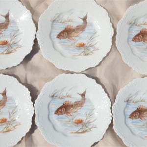 May include: A set of six white porcelain plates with scalloped edges. Each plate features a hand-painted scene of a brown fish swimming in a pond with aquatic plants and a water lily. The plates are arranged on a neutral-toned surface.