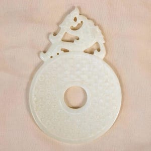 May include: A white jade pendant with a circular design and a central hole. The pendant features a carved dragon motif at the top and a textured surface. The pendant is approximately 10 cm in diameter.