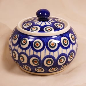 May include: A blue and white ceramic sugar bowl with a lid. The bowl and lid are decorated with a repeating pattern of blue circles with brown and green centres. The lid has a small, round blue knob.