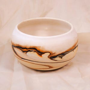 May include: A white ceramic bowl with a rounded shape. The bowl has a marbled design with black, orange, and brown swirls around the exterior. The interior of the bowl is plain white.