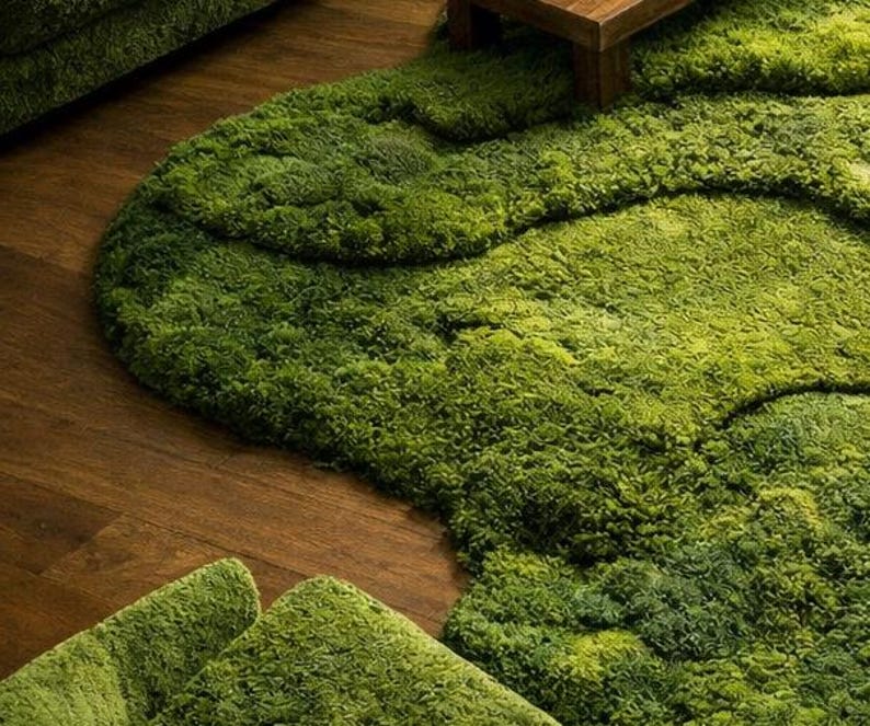 May include: A green, textured rug with an organic, flowing shape, resembling a patch of moss or grass. The rug is placed on a wooden floor, creating a contrast between the natural textures. The rug's design adds a unique, natural element to the room.