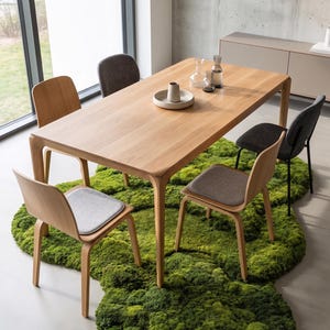 May include: A light wood dining table with rounded edges and six chairs. The chairs have wooden frames and gray seat cushions. The table is set on a green moss-like rug. Glassware and a ceramic piece sit on the table.