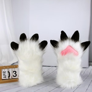 Op de afbeelding: Een paar witte, pluizige poothandschoenen met zwarte klauwen en een roze voetzool. De handschoenen zijn ontworpen om op dierenpoten te lijken. De afbeelding toont de handschoenen tegen een witte achtergrond.