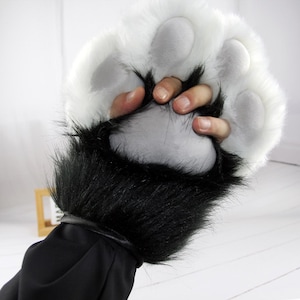 May include: A furry paw-shaped glove with white fur and grey paw pads. The glove has black fur around the wrist and a black cuff. A hand is holding the glove.