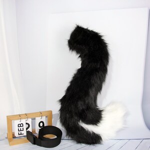 May include: A black and white faux fur tail with a black belt and a wooden calendar displaying "FEB 29". The tail is fluffy and curved, with a black top and a white tip. The belt is black and has a buckle.