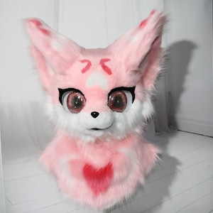 Può includere: Una testa di volpe in peluche rosa e bianca con grandi occhi espressivi e un disegno a forma di cuore sul petto. La testa di volpe ha grandi orecchie a punta con accenti rosa e un naso nero. Gli occhi sono grandi e dettagliati.