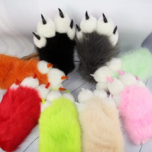 Peut inclure: Une collection de gants en fausse fourrure en forme de pattes colorées avec des griffes noires. Les gants sont de différentes couleurs, dont le noir, le gris, l'orange, le rouge, le vert, le rose et le beige. Les pattes ont des coussinets blancs.