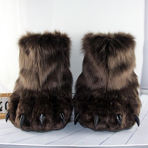 Può includere: Un paio di pantofole a forma di zampa d'orso in finta pelliccia marrone. Le pantofole hanno un design realistico con artigli marrone scuro e una consistenza soffice. Le pantofole sono progettate per assomigliare a zampe d'orso.