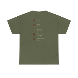 Op de afbeelding: Olijfgroen t-shirt met het woord "GRIND" verticaal gedrukt in een roestkleurig lettertype. Elke letter van "GRIND" komt overeen met een woord: God, Rust, Veerkracht, Integriteit, Intentie, Noodzakelijke Actie, Toewijding, Drive.