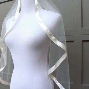 May include: A sheer white bridal veil with a satin ribbon trim. The veil is draped over a white mannequin, showcasing its delicate texture and design. The ribbon trim adds a touch of elegance to the wedding accessory.