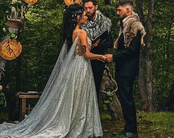 May include: A wedding ceremony featuring a person in a floor-length silver sequined gown with a long cape, a person in a black suit with a fur shoulder wrap, and an officiant in a black suit with a fur collar, standing before a wooden arch with runes.