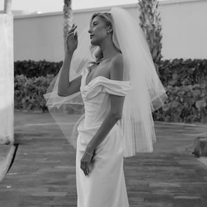 Op de afbeelding: Een bruid in een witte, off-shoulder bruidsjurk met een lange sluier. Ze staat op een geplaveide weg met palmbomen op de achtergrond.
