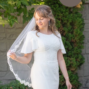 Puede incluir: Una novia con un vestido de novia blanco con detalles de encaje y un velo blanco. Está de pie frente a una pared de ladrillo y vegetación verde.