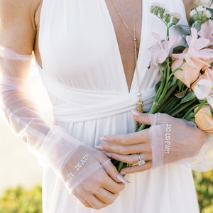 May include: A white wedding dress with a plunging neckline. The bride is wearing sheer gloves with the words "TIL DEATH" and "DO US PART" embroidered on them. She holds a bouquet of flowers and wears a gold necklace.