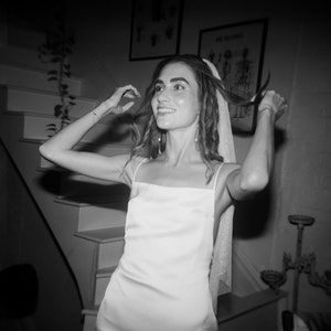 Pode incluir: Uma mulher com um vestido branco de cetim e véu, sorrindo e ajustando o cabelo.