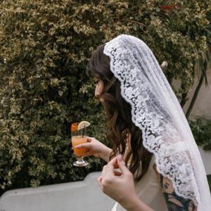 K&ouml;nnte beinhalten: Eine Frau in einem wei&szlig;en Spitzenkleid und einem wei&szlig;en Schleier h&auml;lt ein Glas Orangensaft mit einer Zitronenscheibe. Sie hat ein Tattoo auf ihrem Arm.
