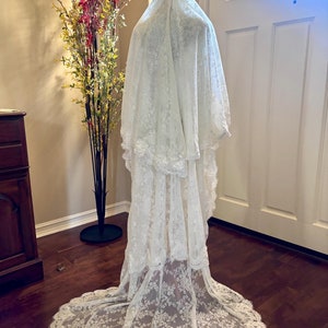 Puede incluir: Un velo de novia de encaje blanco con un borde festoneado. El velo está drapeado sobre un maniquí y tiene una cola larga.