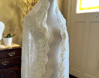 May include: A white lace shawl with a scalloped edge. The shawl is draped over a white mannequin.